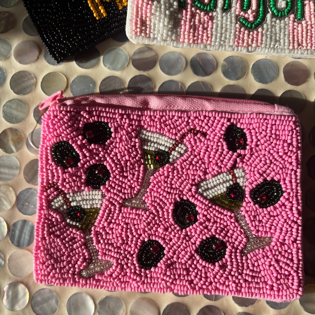 A pink beaded zippered pouch featuring a pattern of martini glasses and black olives, laid flat on a circular mother-of-pearl tile surface.