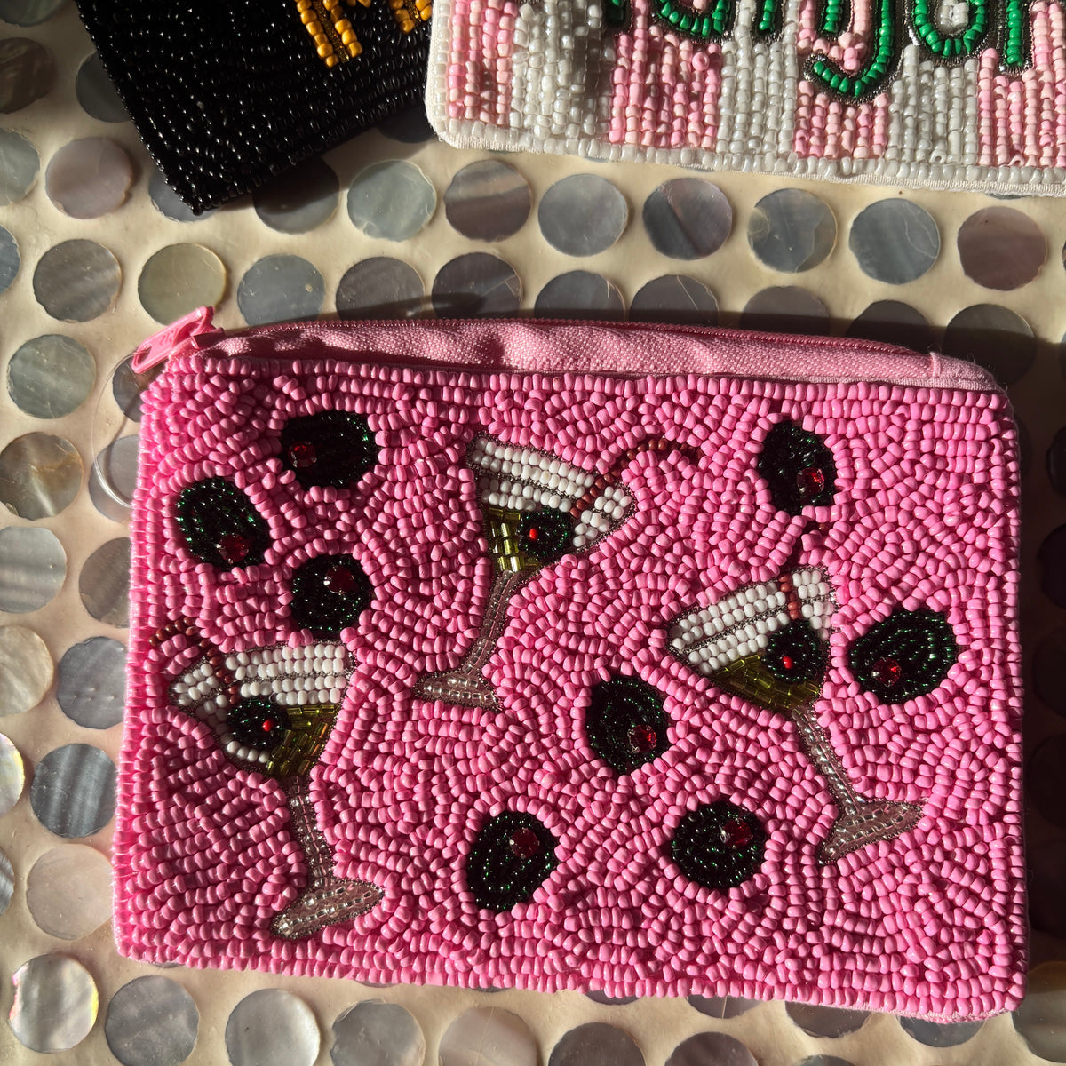 A pink beaded zippered pouch featuring a pattern of martini glasses and black olives, laid flat on a circular mother-of-pearl tile surface.