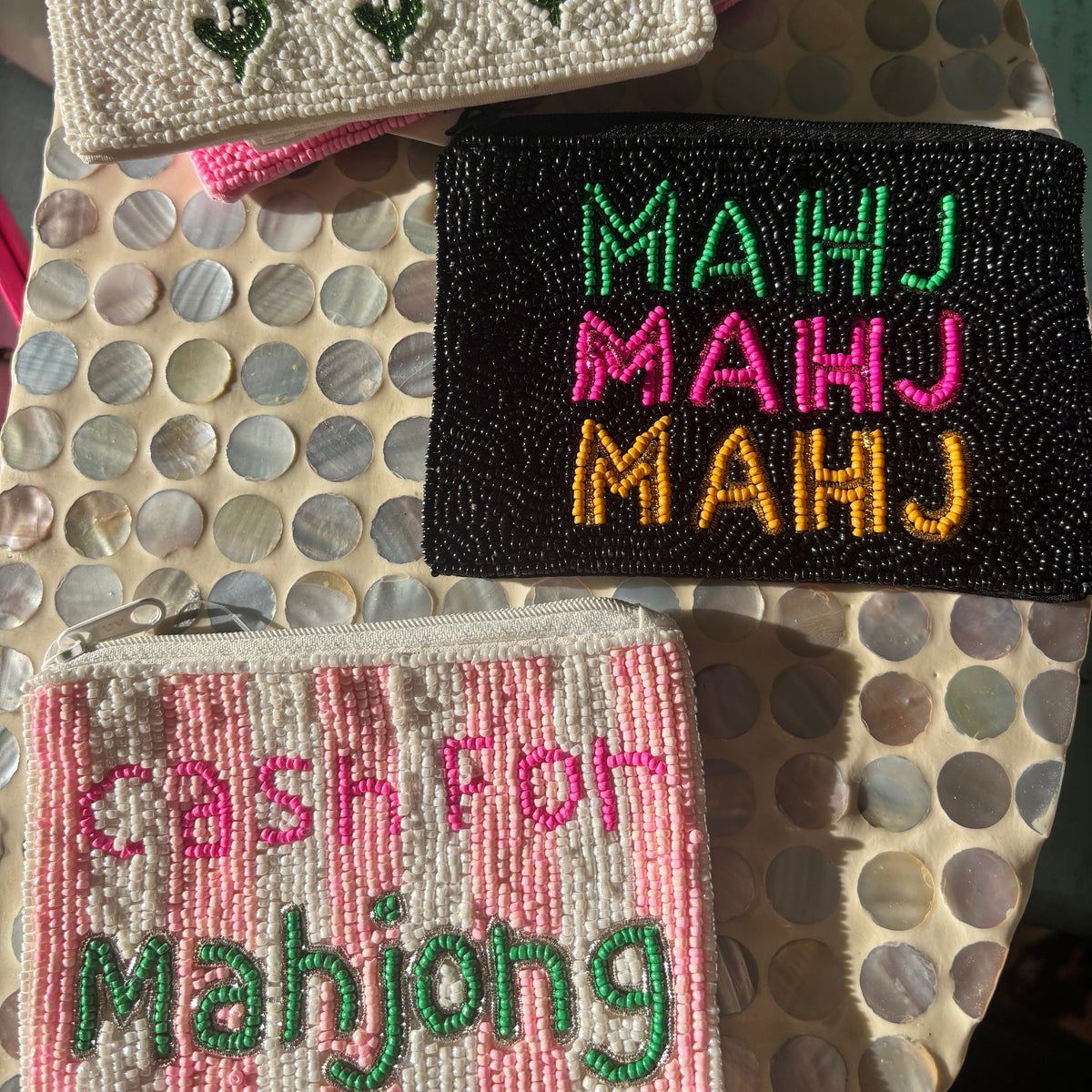 Three beaded coin purses for Mahjong displayed on a mother-of-pearl table. One cream purse has green dollar signs and maroon text reading 'MAHJOUT $$$'.