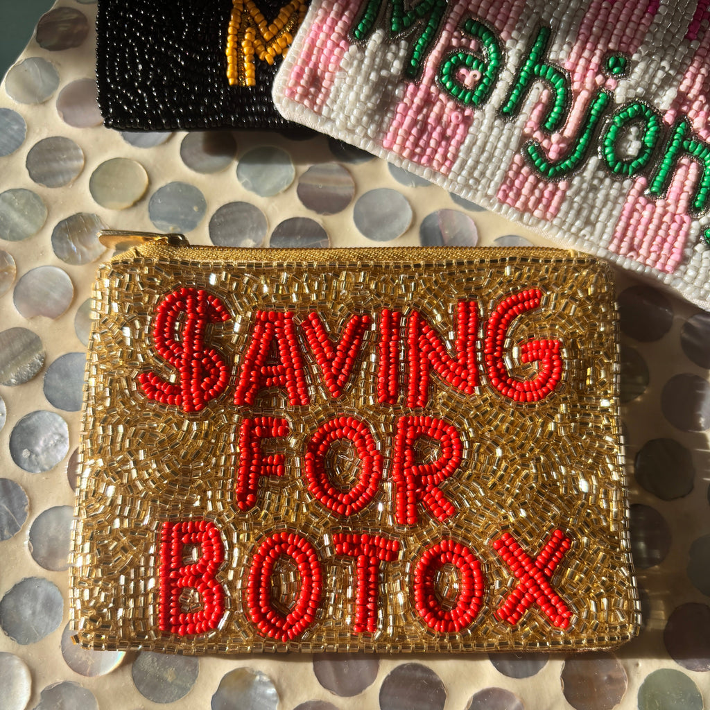 A gold beaded zip pouch with red bead lettering that reads 'SAVING FOR BOTOX' in all caps, displayed on a mother-of-pearl patterned surface with other beaded accessories partially visible in the background.