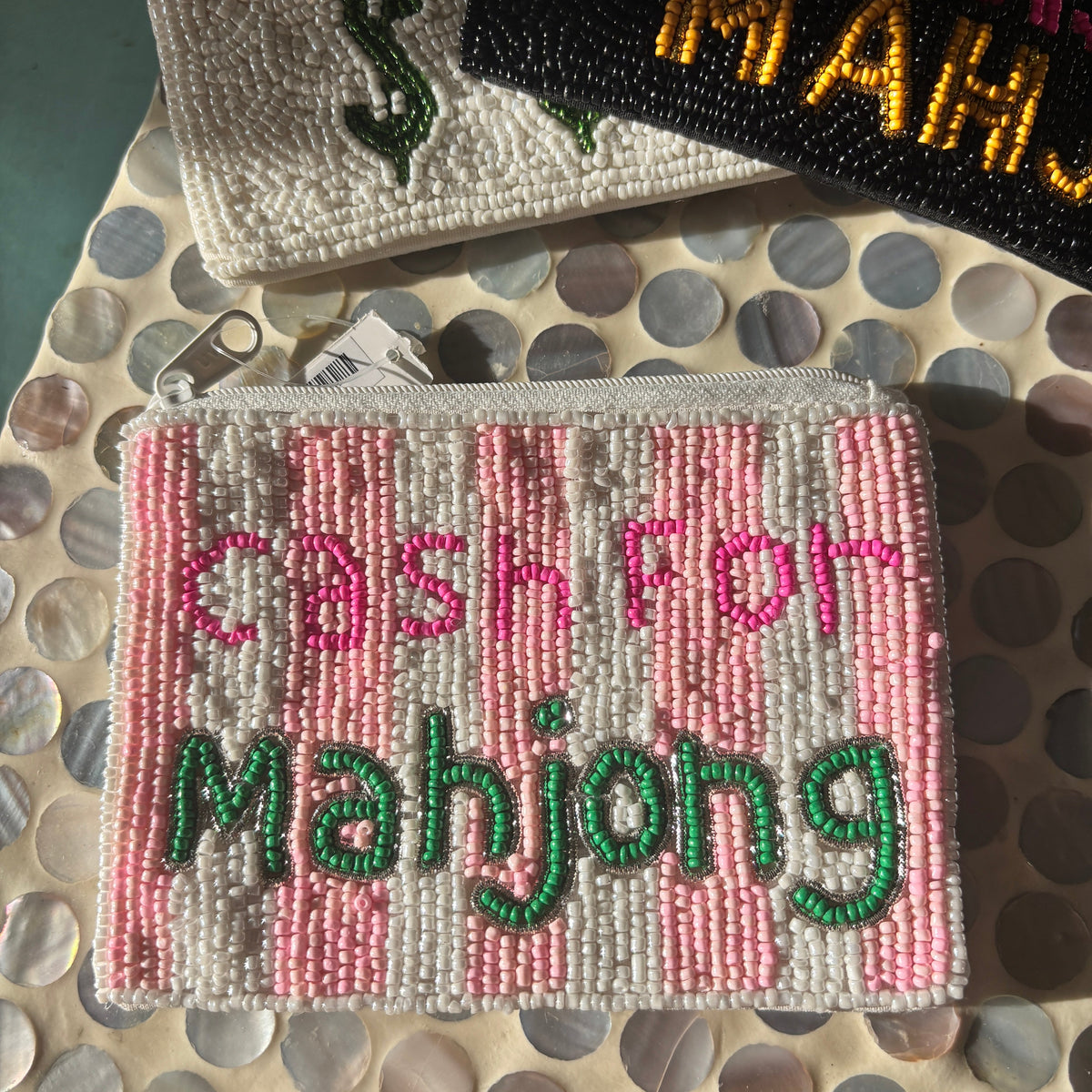A rectangular beaded coin purse with pink and white stripes and the words 'CASH FOR mahjong' in pink and green beads, displayed on a mother-of-pearl tile surface with other beaded purses partially visible behind it.