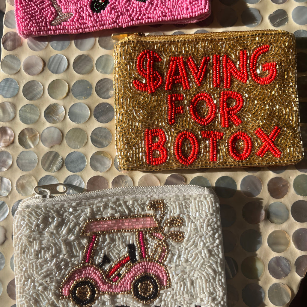 Three beaded zip pouches displayed on a circular shell-patterned surface. The top pouch is bright pink with black bead details.