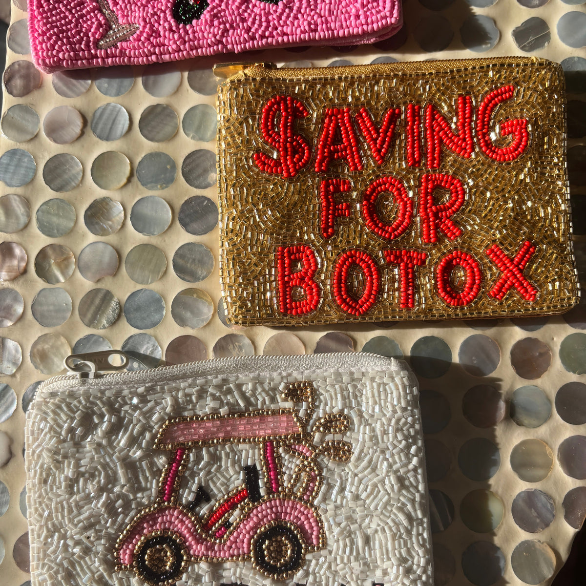 Three beaded zip pouches displayed on a circular shell-patterned surface. The top pouch is bright pink with black bead details.
