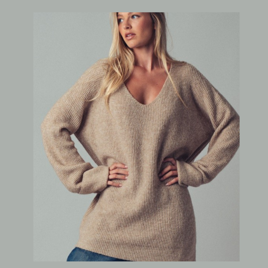 A woman wearing a beige ribbed knit sweater with a wide V-neck and long sleeves, posed against a solid gray background.