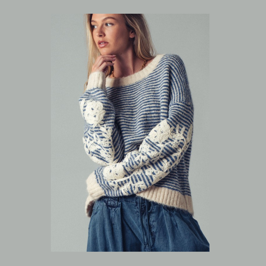 A woman wearing a blue and cream knit sweater with textured patches on the sleeves, paired with blue jeans, set against a solid gray background.