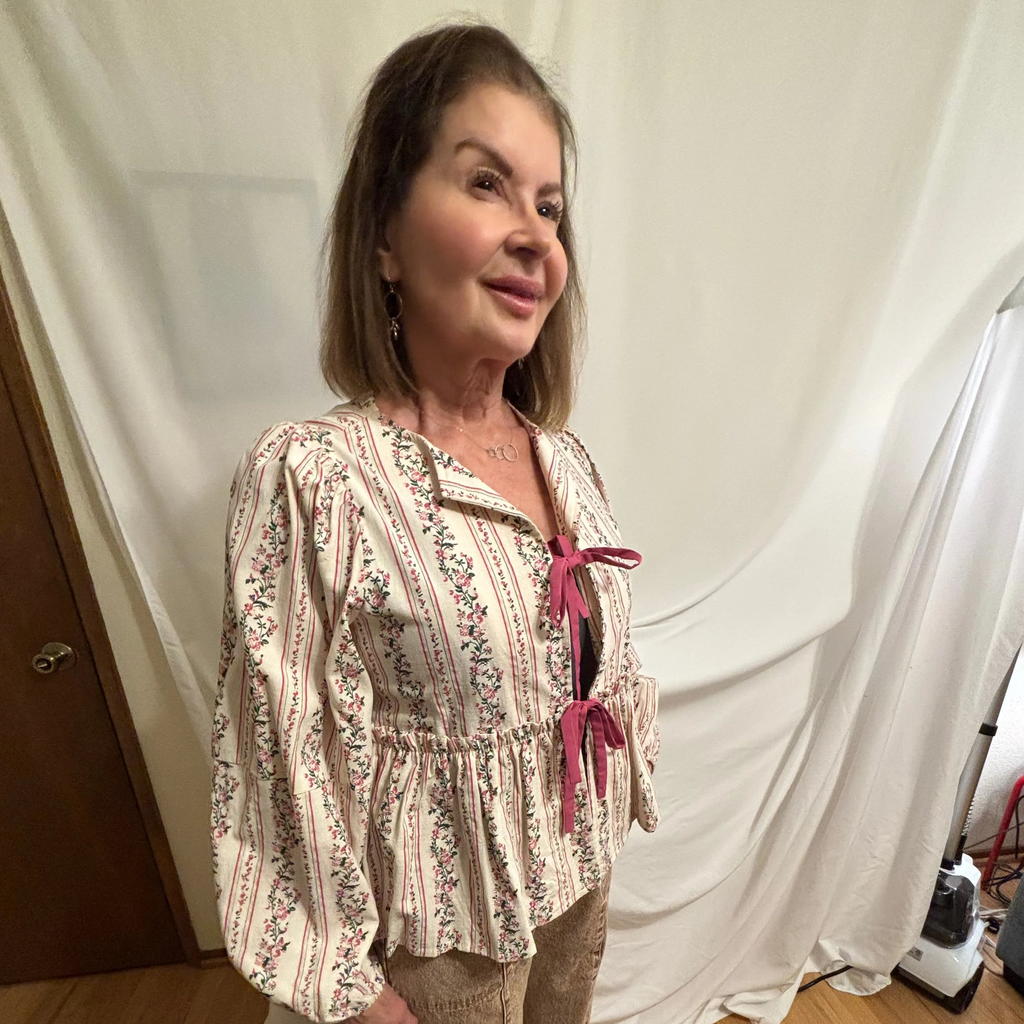 Woman wearing a patterned blouse and beige pants standing in a room with white curtains.