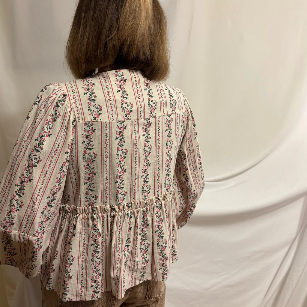 Person wearing a floral blouse and brown pants standing against a white curtain.