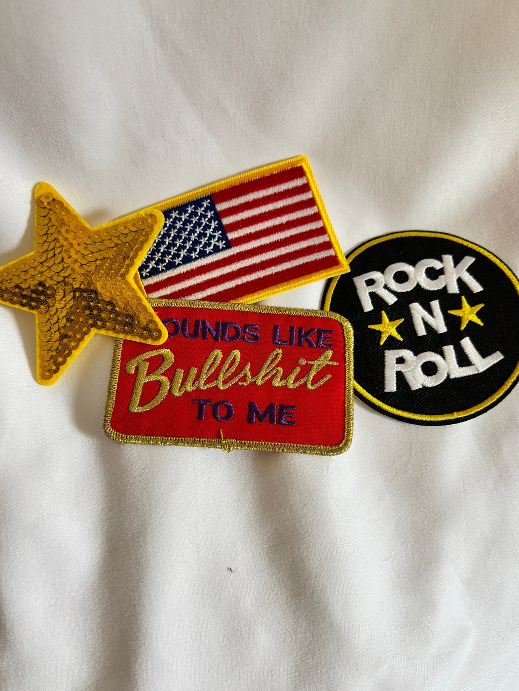 Collection of patches including 'Rock N Roll', American flag, and 'Bullshit to Me' on a white fabric background.