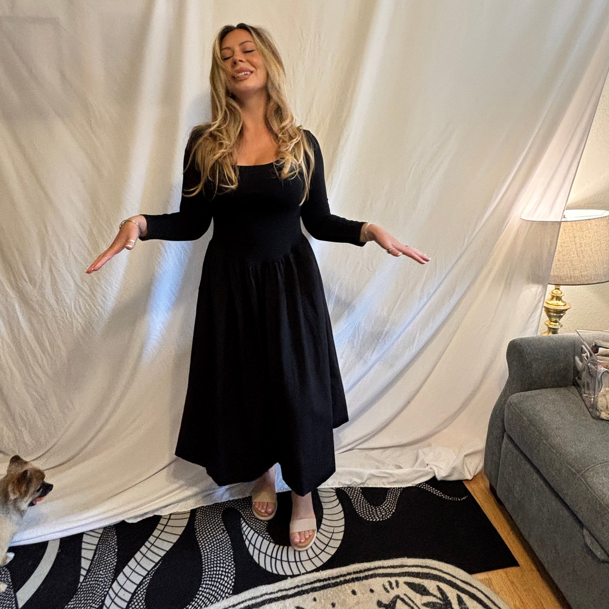 A woman wearing a black long-sleeve midi dress stands with her arms outstretched in front of a draped white fabric backdrop. A patterned rug, a gray sofa, and a lamp are visible in the room setting.