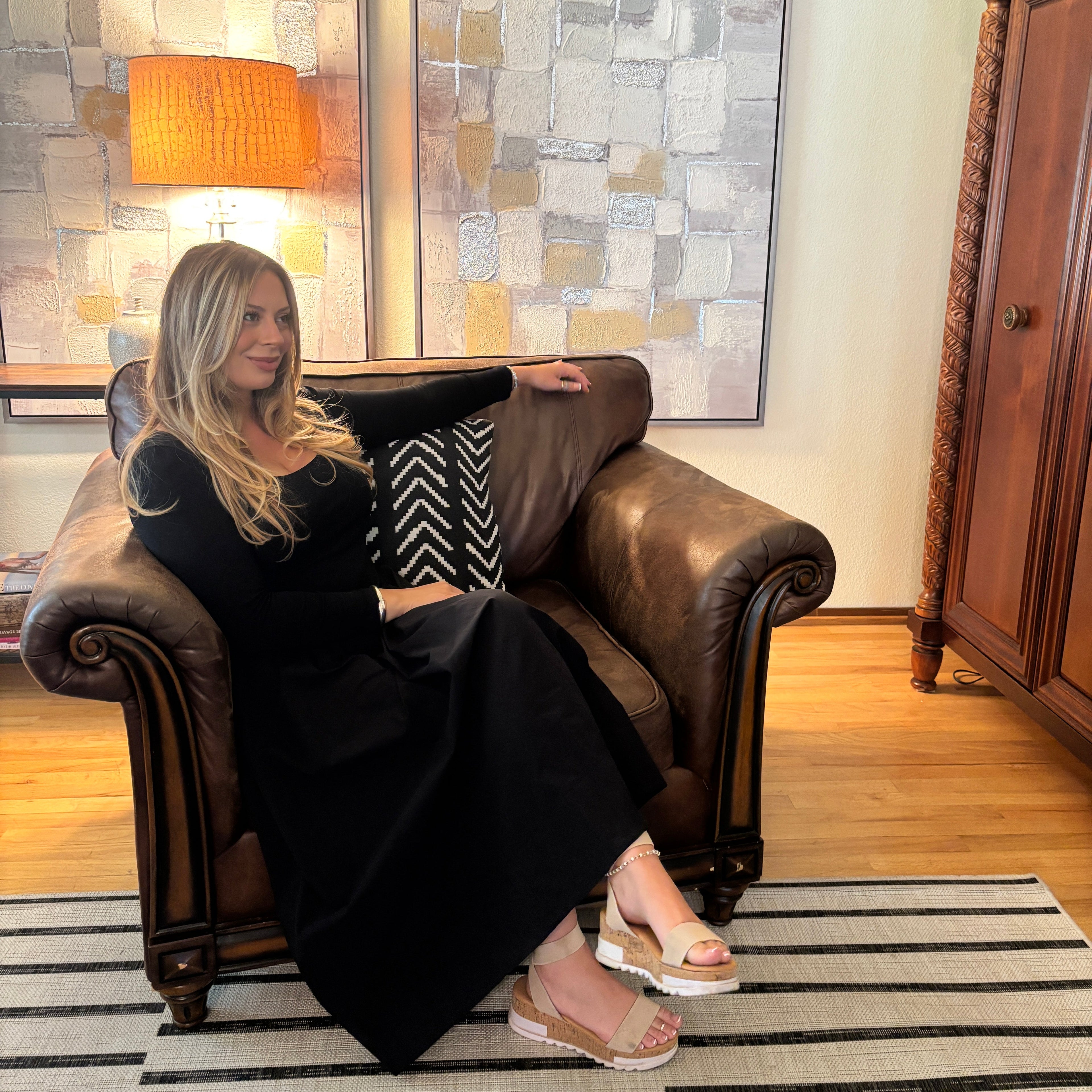 A woman with blonde hair sits in a brown leather armchair in a living room setting, wearing a long black dress and beige platform sandals. The room features two framed abstract paintings on the wall, a wooden floor, and a striped rug.