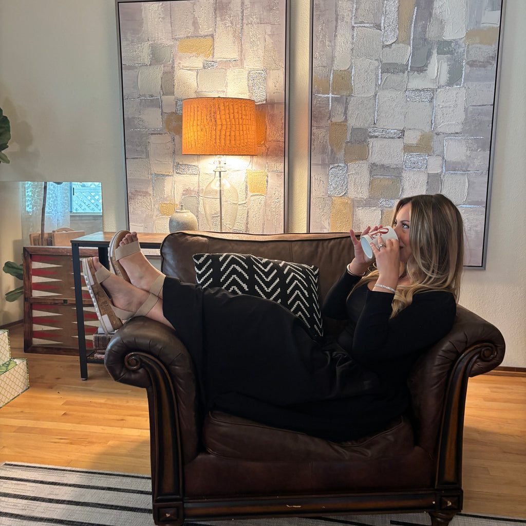 A woman reclining in a dark brown leather armchair, holding a mug and looking at a phone. She is wearing black pants and a black top.