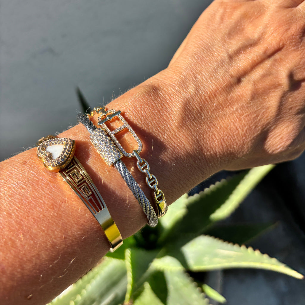 A close-up of a person's wrist wearing multiple bracelets, including a gold bangle with a red geometric pattern, a gold heart-shaped bezel bracelet, a silver cable chain bracelet, and a silver link bracelet with a gold rectangular accent. A green succulent is visible in the background against a gray wall.