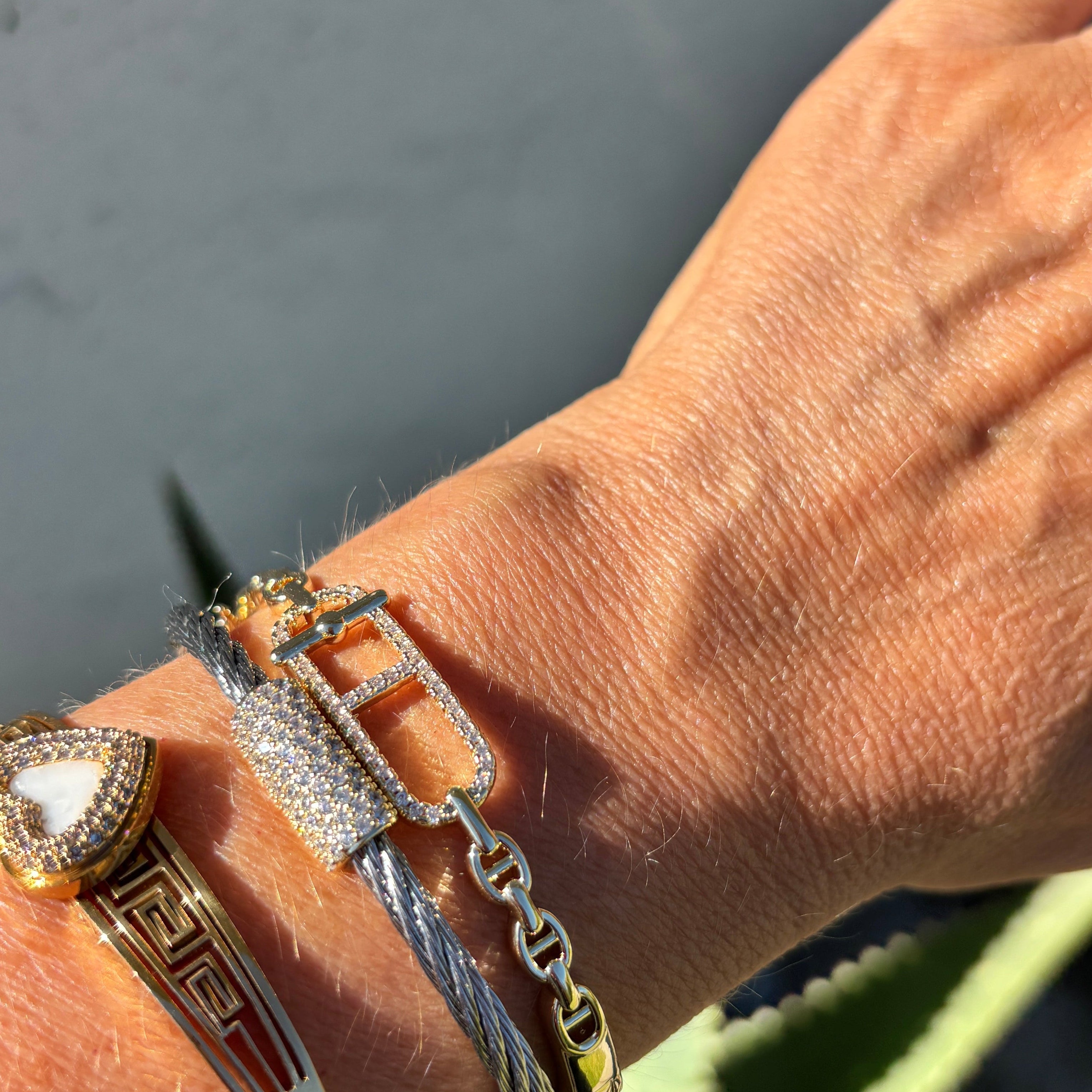 A close-up of a person's wrist wearing multiple bracelets, including a gold bangle with a red geometric pattern, a gold heart-shaped bezel bracelet, a silver cable chain bracelet, and a silver link bracelet with a gold rectangular accent. A green succulent is visible in the background against a gray wall.
