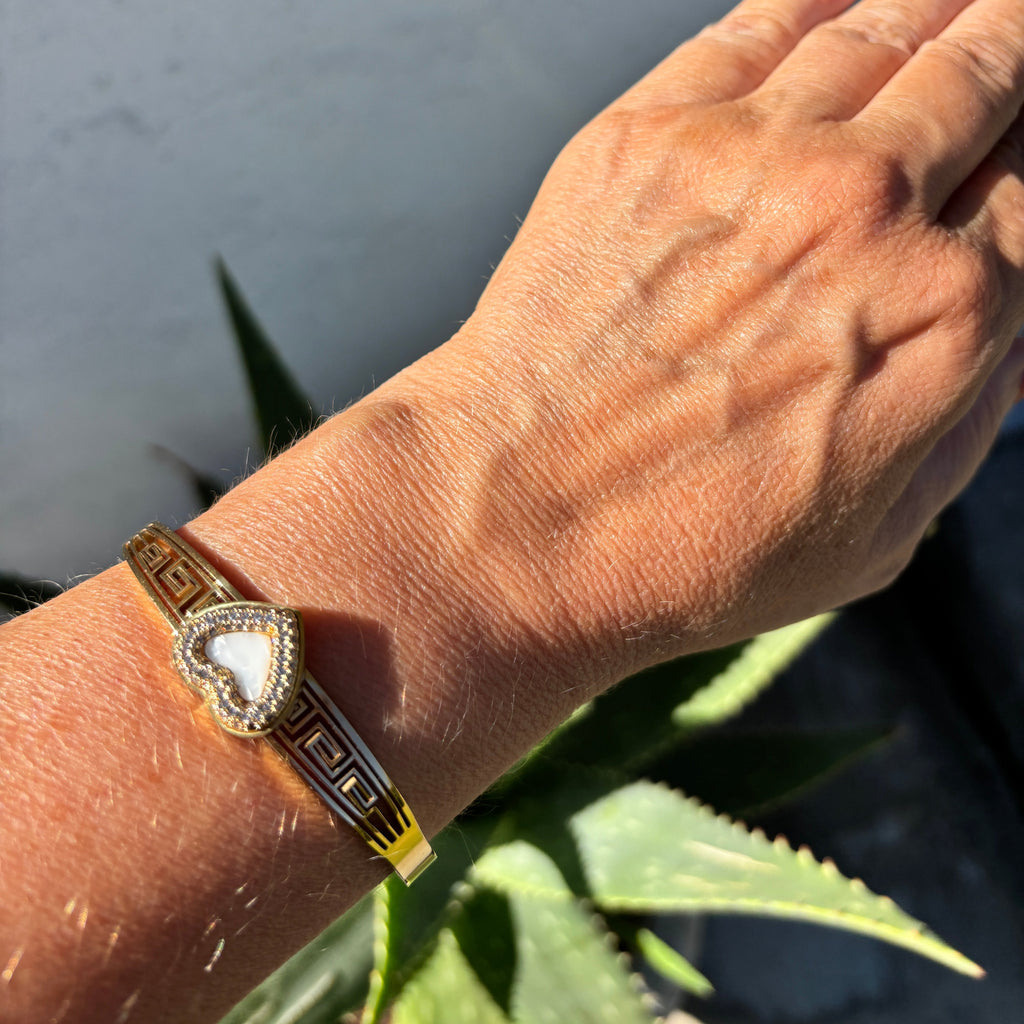 A gold and silver bangle bracelet with a heart-shaped crystal pendant, worn on a person's wrist. The person's hand has red nail polish, and green aloe vera leaves are visible in the background against a gray wall.