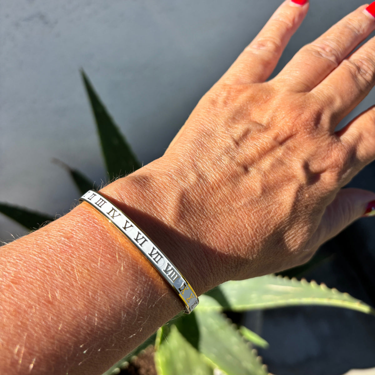 A close-up of a person's wrist wearing a silver metal band bracelet engraved with the Roman numerals I, V, VI, VII, VIII, IX, X, XI, XII, XIII, XIV, XV, XVI, XVII, XVIII, XIX, XX, XXI, and XXII.
