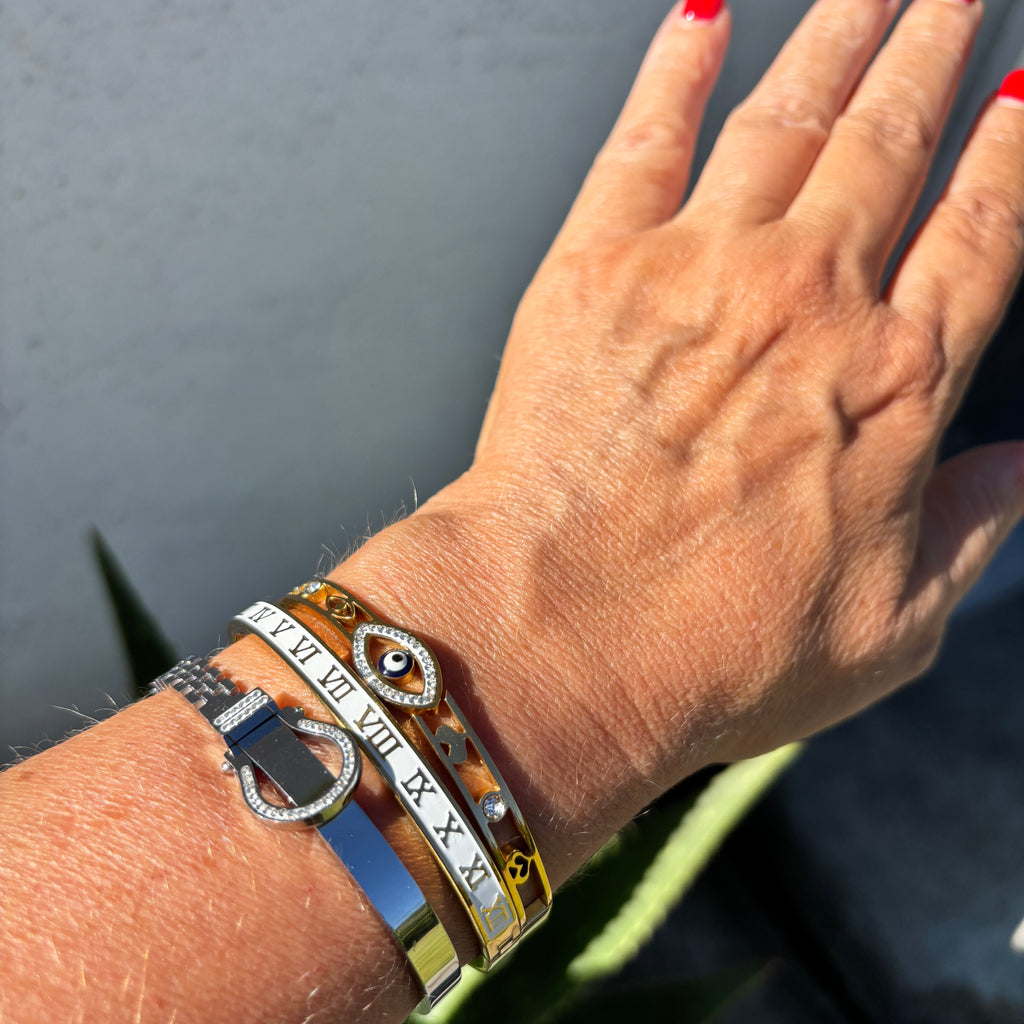A close-up of a person's wrist wearing multiple bracelets, including a silver-tone bangle with a horsebit motif, a white bangle with Roman numerals, and a gold bangle with a circular emblem, against a dark gray background with green leaves at the bottom.