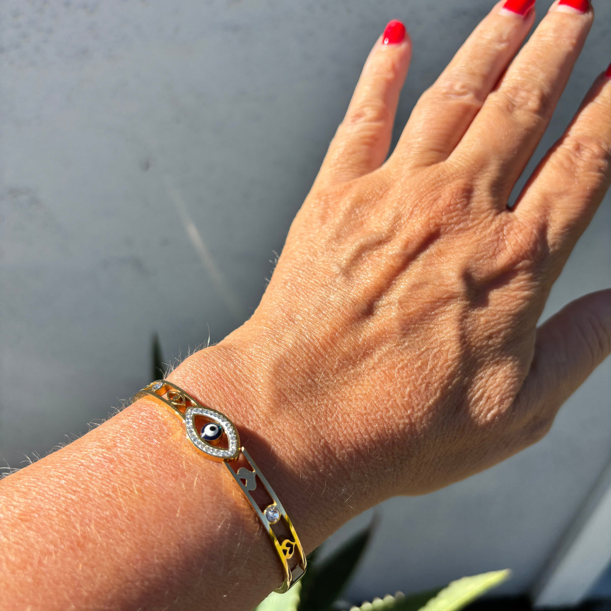 A gold-toned bangle bracelet with a central evil eye motif and small heart details, worn on a person's wrist. The person's hand has red nail polish, and the background is a neutral gray wall with a hint of green foliage.