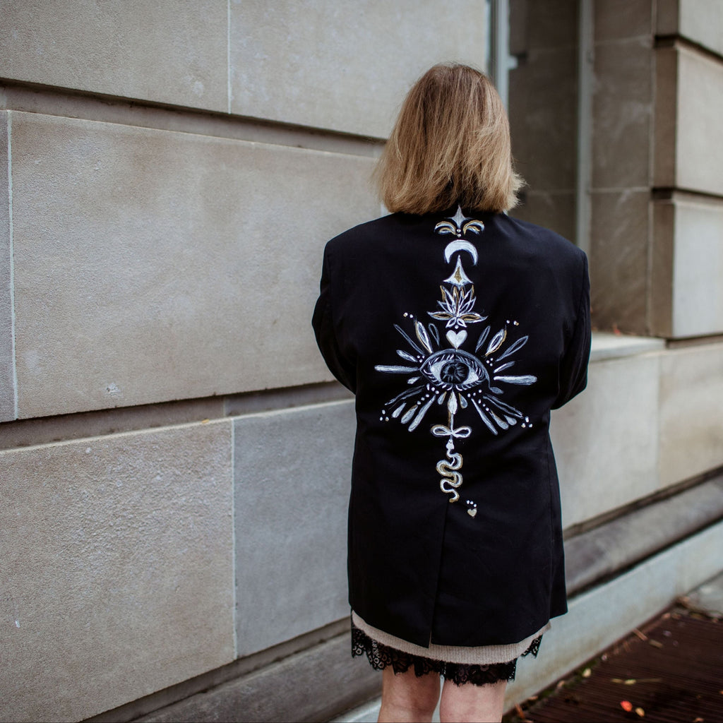Person wearing a black jacket with a detailed design on the back, standing against a stone building.