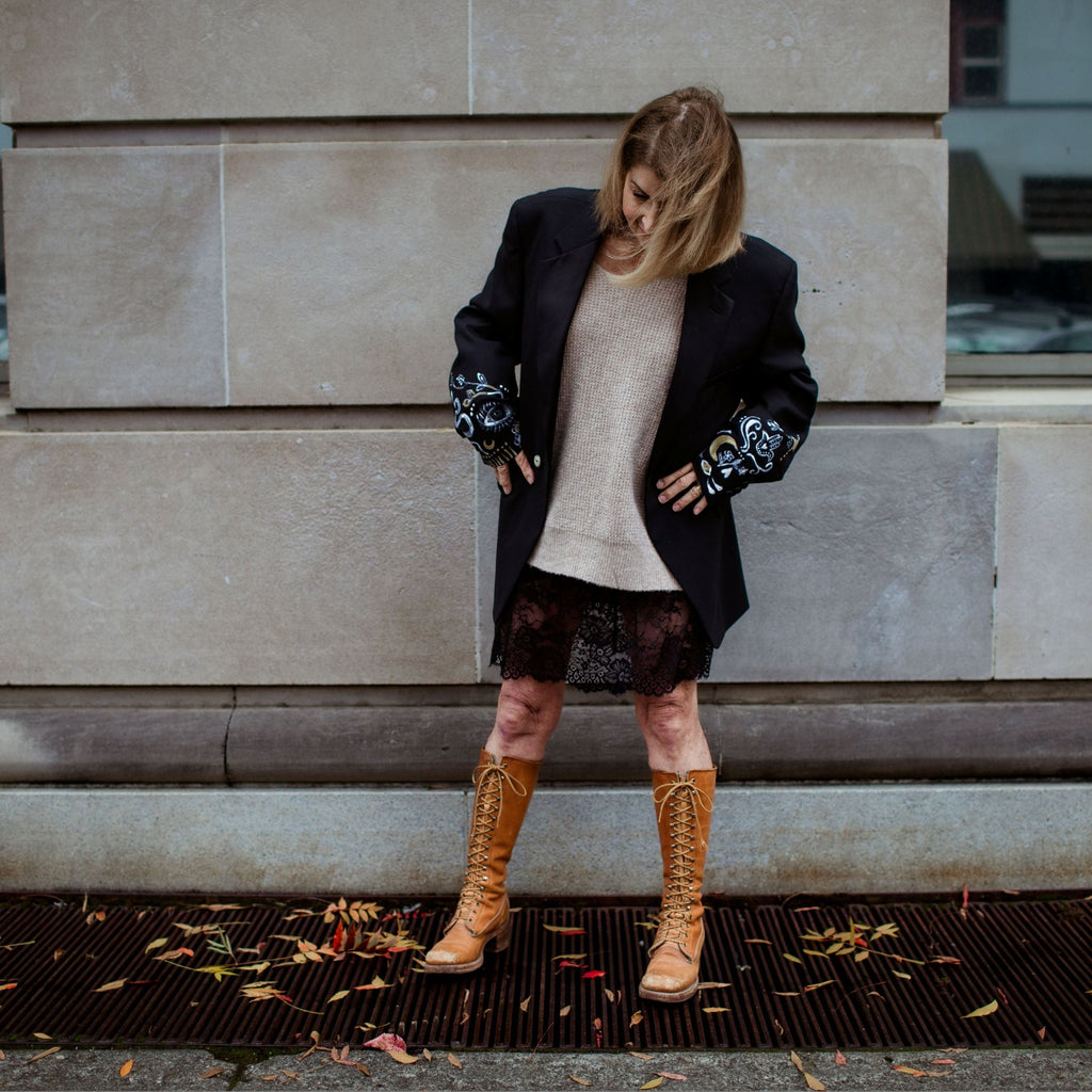 Woman standing on a city street wearing a dark coat, light sweater, and patterned pants.