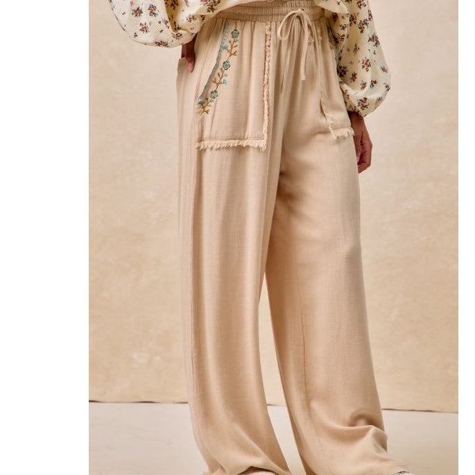 Person wearing beige wide-leg pants with floral embroidery and a floral blouse against a beige background