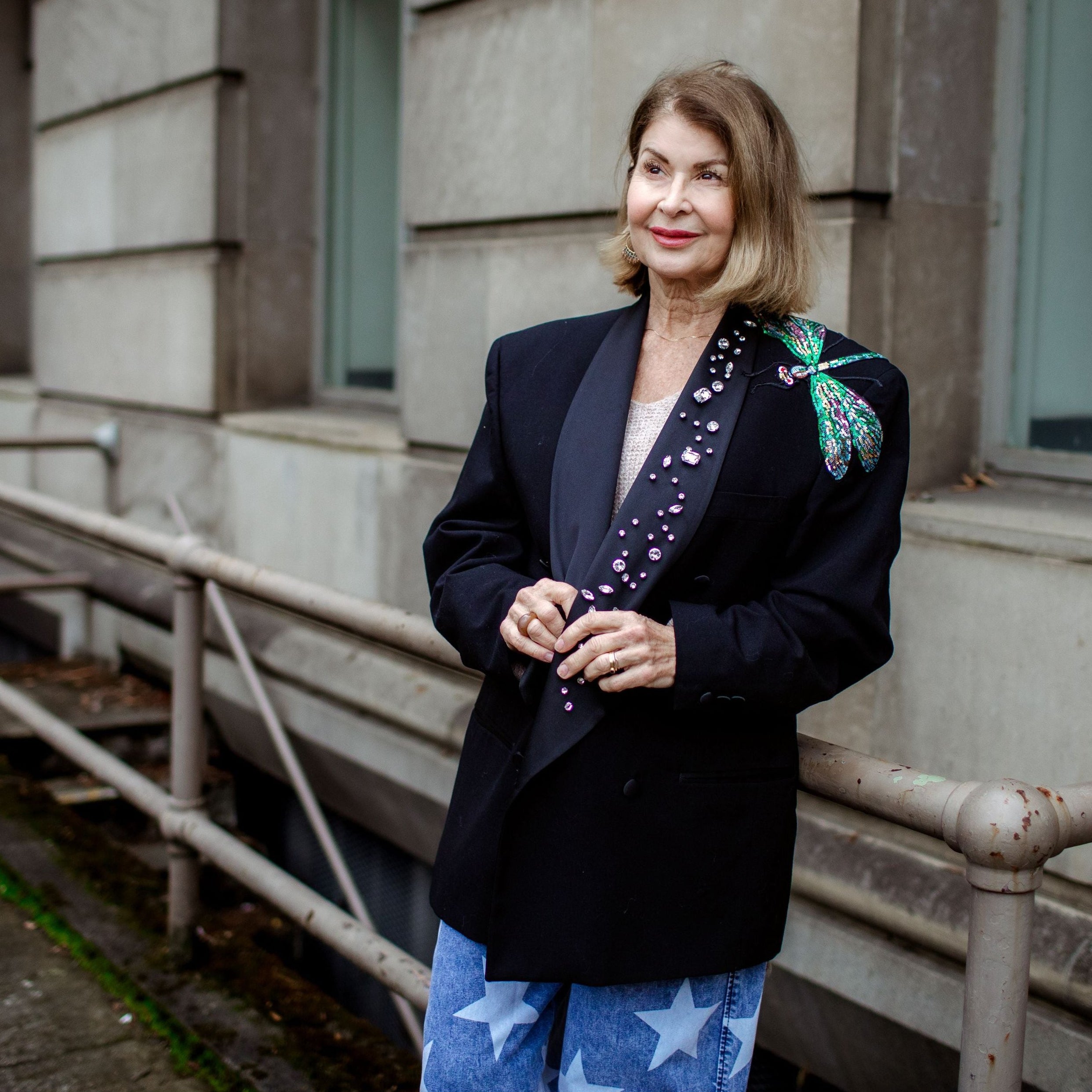 Woman standing outside a building wearing a black blazer with floral embroidery and blue pants with star patterns.