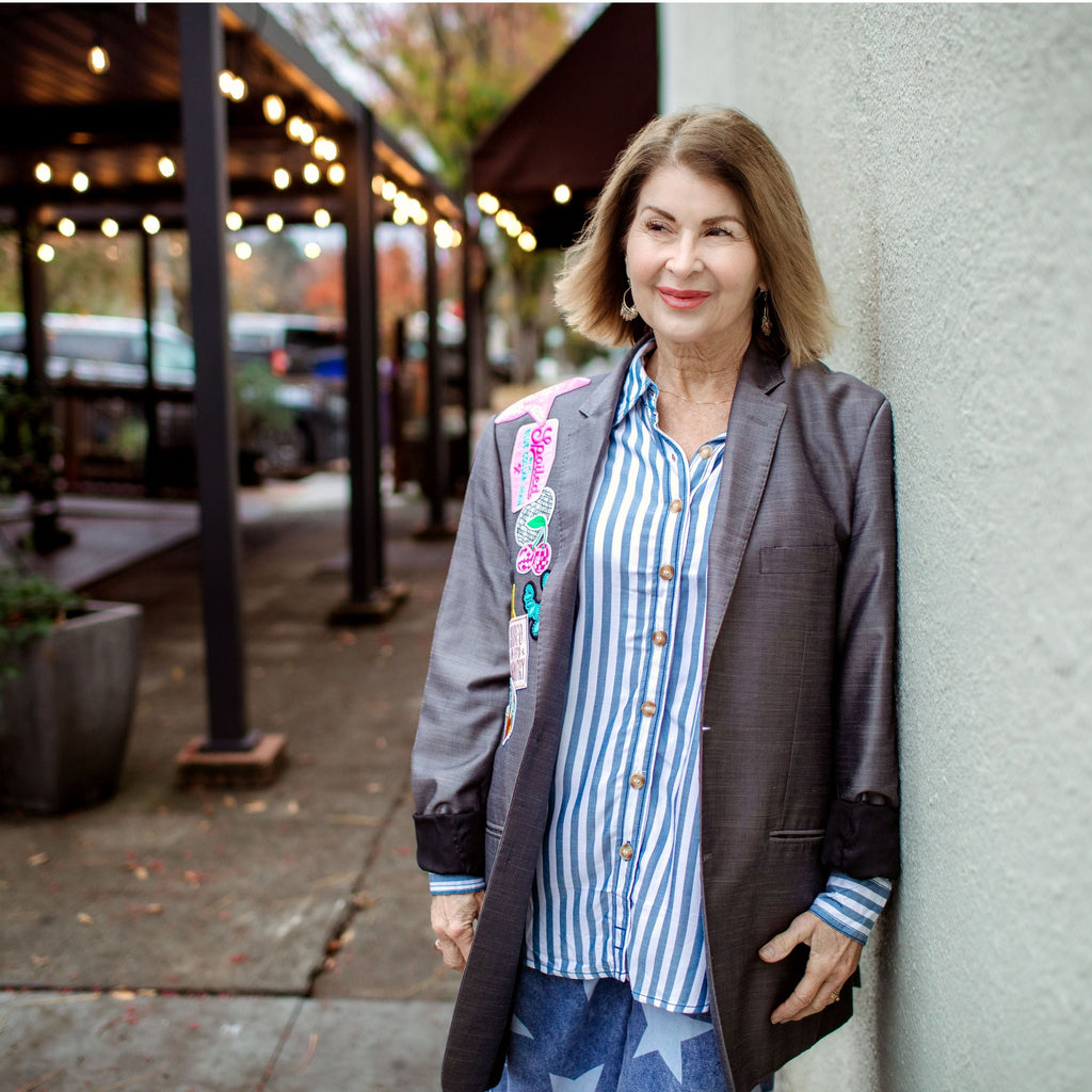 Woman standing against a wall in an outdoor setting with plants and a building in the background.