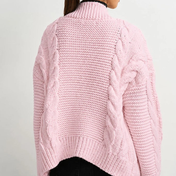 A woman shown from the back wearing a light pink chunky cable-knit sweater with a mock neck, paired with black pants, against a plain light gray background.